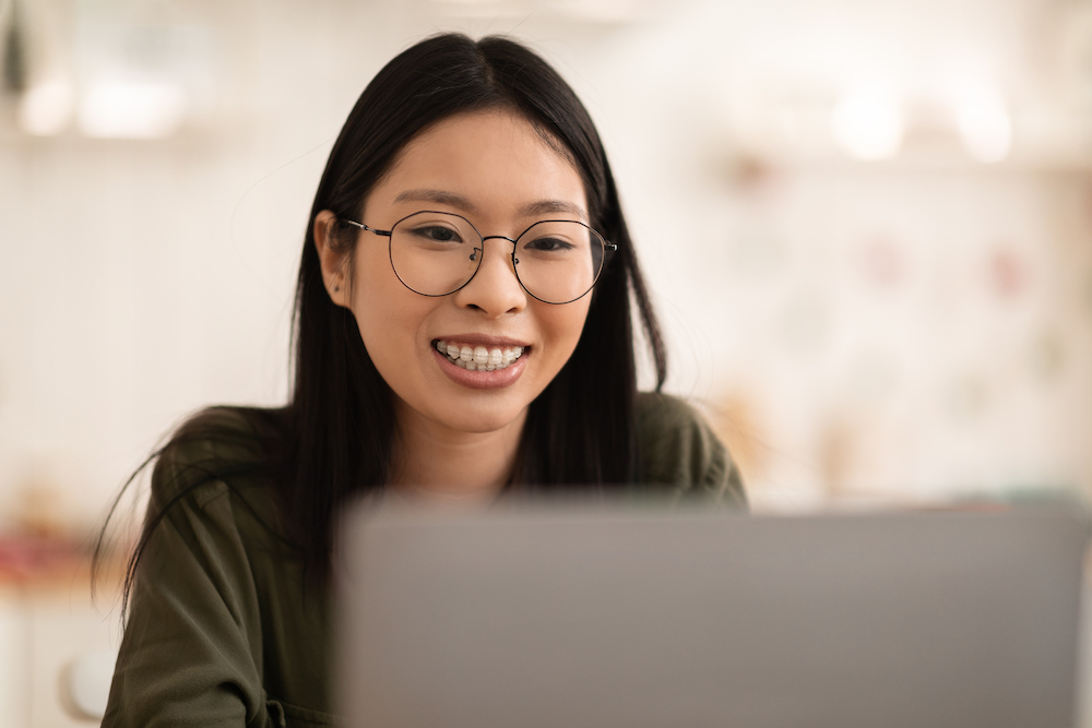 Closeup photo of happy cheerful young asian chinese woman wearing eyeglasses using laptop at kitchen, looking at computer screen and smiling, copy space. Freelance, remote job concept